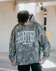 Camo ROOTED hoodie streetwear design with bold lettering and ‘Colossians 3:3’ Scripture detail worn by model.