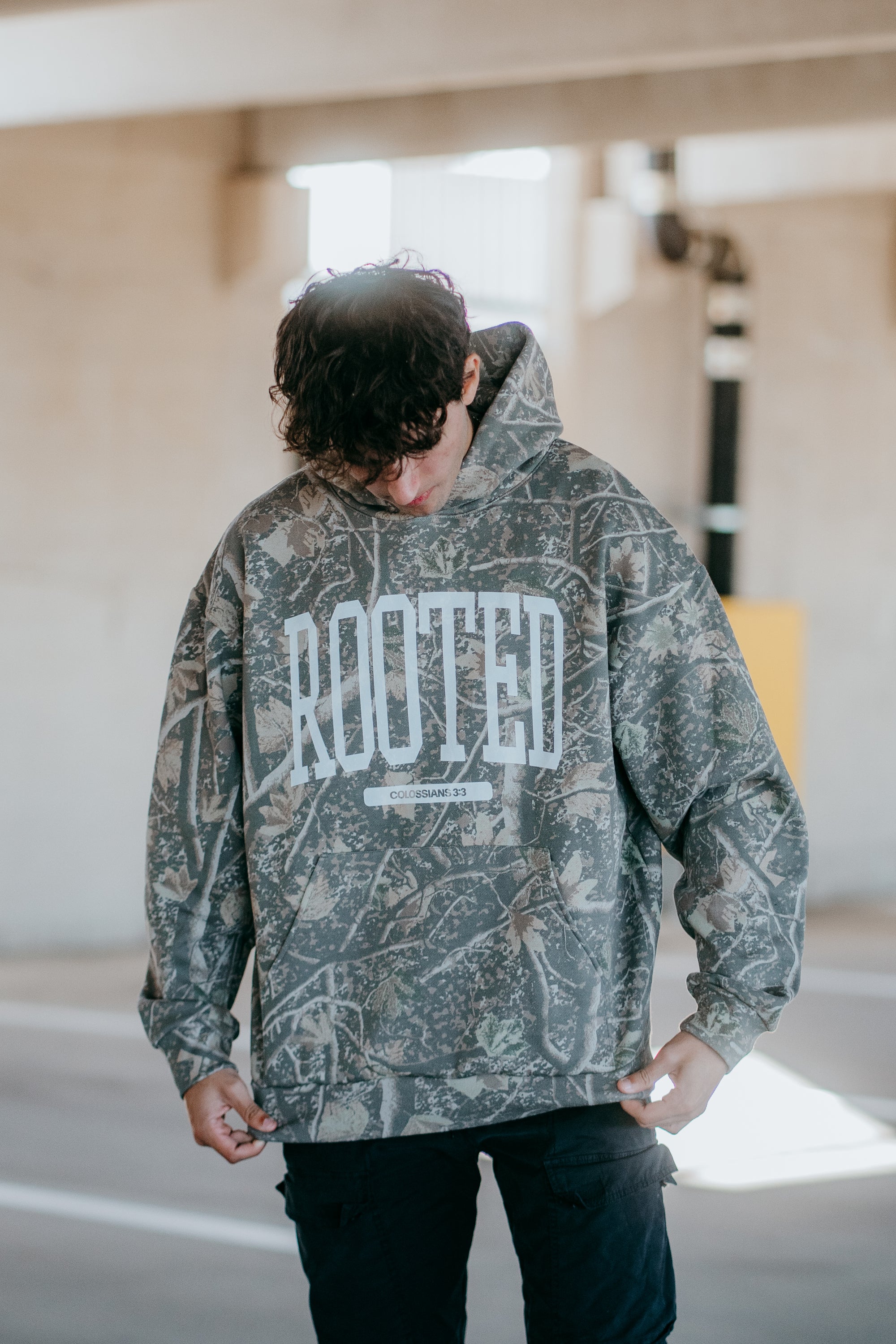 Camo ROOTED hoodie streetwear design with bold lettering and ‘Colossians 3:3’ Scripture detail worn by model.